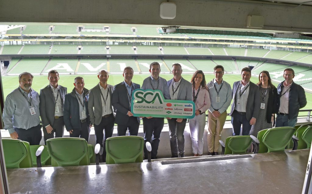 John Paul Construction team members and founding industry partners holding a large green launch sign for the Sustainability Common Assessment (SCA) inside a stadium box overlooking the pitch at the Aviva Stadium, Dublin.